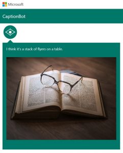 Photo shows Captionbot.ai from Microsoft which has been shown a photo of a pair of glasses resting on an open book, which it believes is a “stack of flyers on a table”.