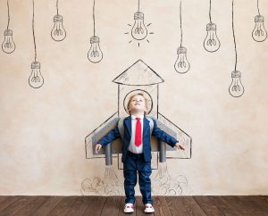 A young boy in a suit stands against a wall looking skywards with his arms out-stretched. Hand drawn, in black and white, around him is the shape of a rocket and various lightbulbs hang down overhead with the one immediately above him apparently lit up.