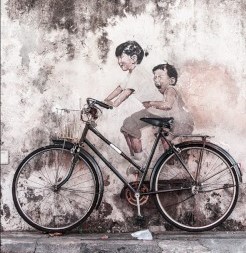 A realist street art wall mural of two life-size children positioned to appear as if they are riding an actual bicycle that is parked on a city sidewalk.