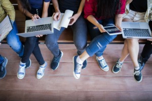 Photograph of students, taken from the neck down in a seted position. Between them they have a laptop, a book and a tablet open 