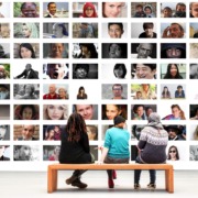 3 people are sat on a bench, backs to the camera, looking at a wall of photographs of many different faces, used here to depict the large reach of The DAISY Consortium.