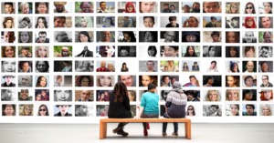 3 people are sat on a bench, backs to the camera, looking at a wall of photographs of many different faces,  used here to depict the large reach of The DAISY Consortium.