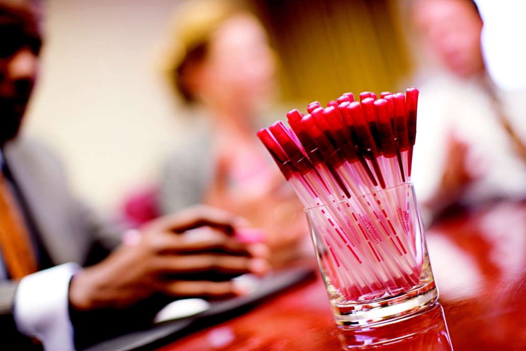 a glass filled with identical red biros on a table with blurred figures in the background denoting a meeting, seminar or conference setting
