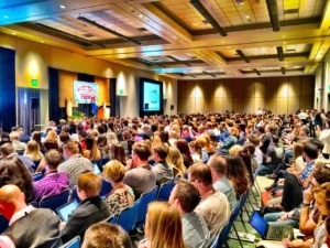 Photograph of an audience at a conference taken from the side of room and also showing the stage