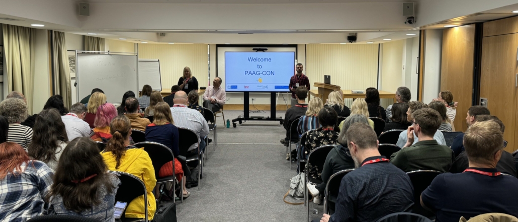 photo showing Stacy Scott welcoming a room full of delegates to PAAGCon, alongside Simon Mellins and Richard Orme