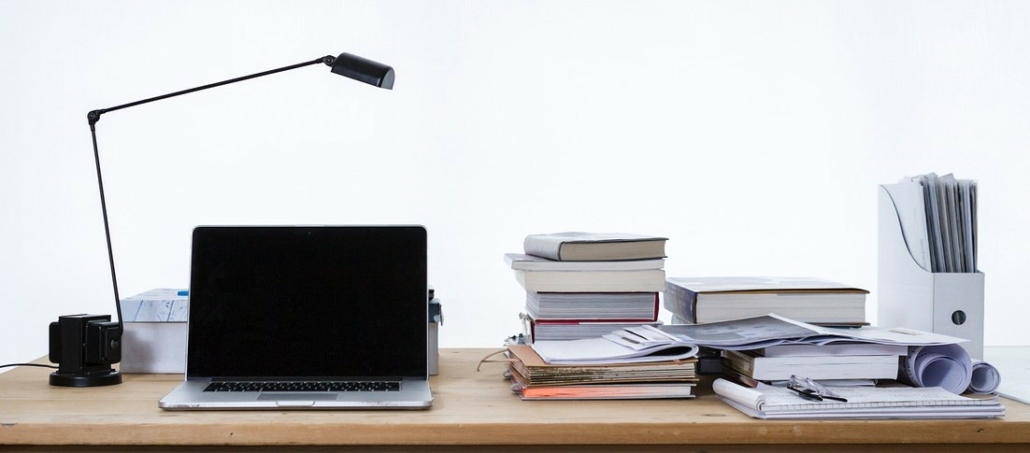 A laptop positioned on the left hand side of a desk full of papers and books. A lamp is placed above the laptop which is the main focus of the image.