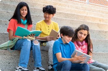 A group of middle school students. They are sitting on concrete steps and reading from books and mobile devices.